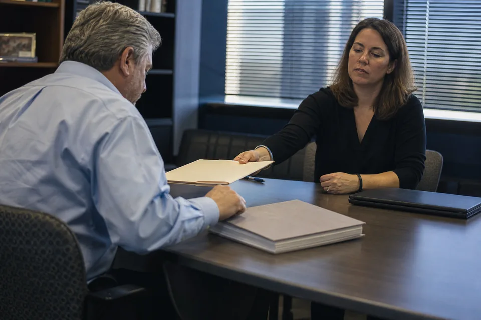 Contracts manager and a program manager at a small federal contractor reviewing a binder of subcontracts in a conference room